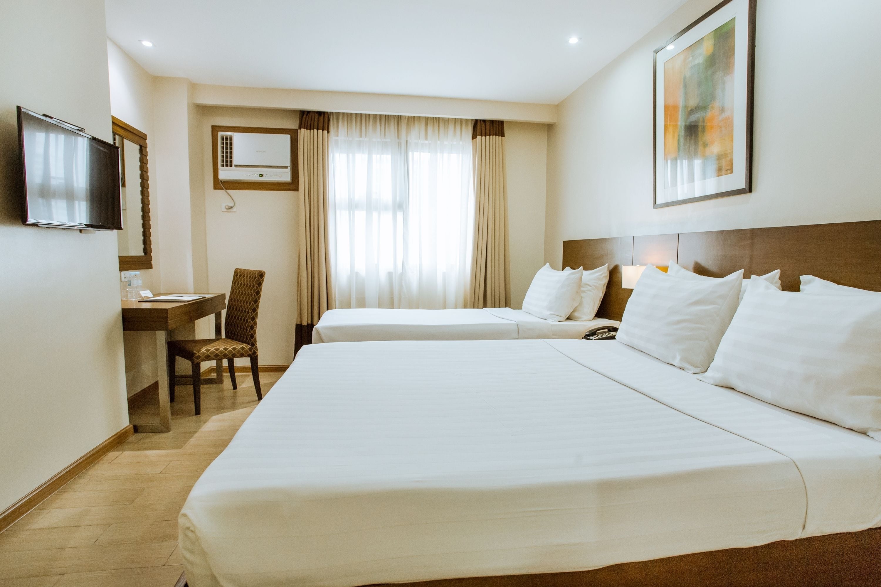 A hotel room with two beds, a desk, a chair, a television, and an air conditioning unit.