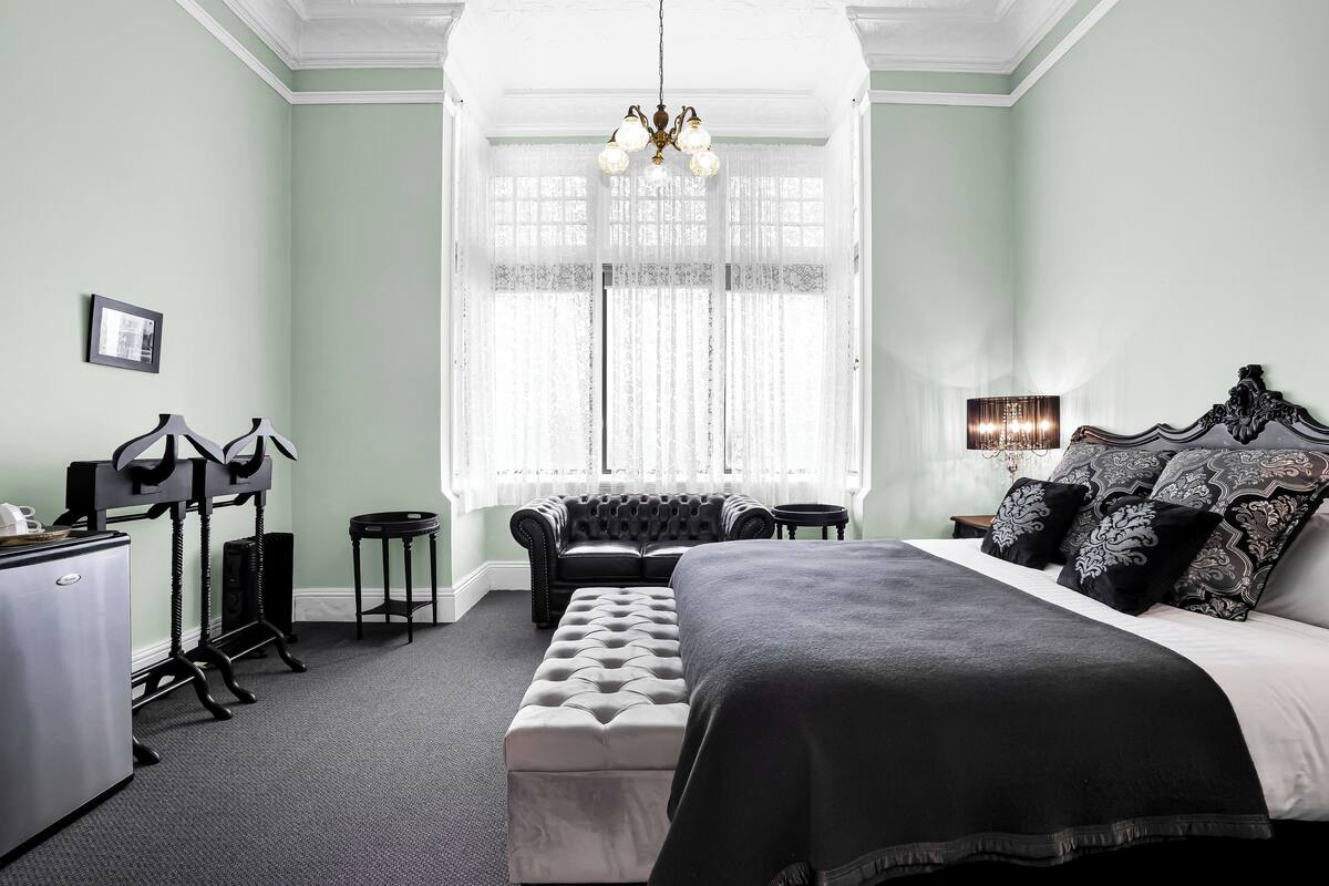 Ein Schlafzimmer mit einem großen Bett, einer Bank, einem Schminktisch, einem Spiegel, einem Fenster mit Vorhängen und einer Chandelier.