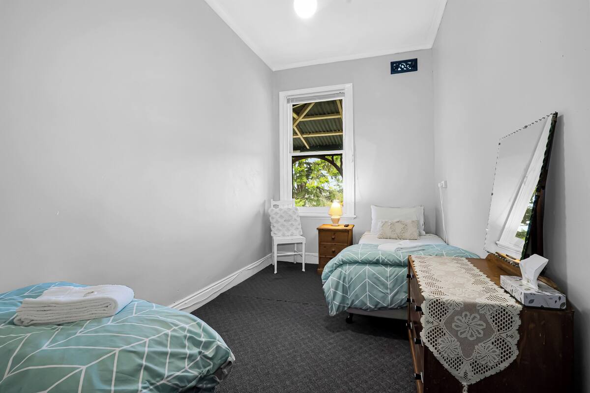 Ein Schlafzimmer mit zwei Betten, einem Sessel, einem kleinen Tisch mit Lampe und einem Fenster mit Blick auf Grünflächen.