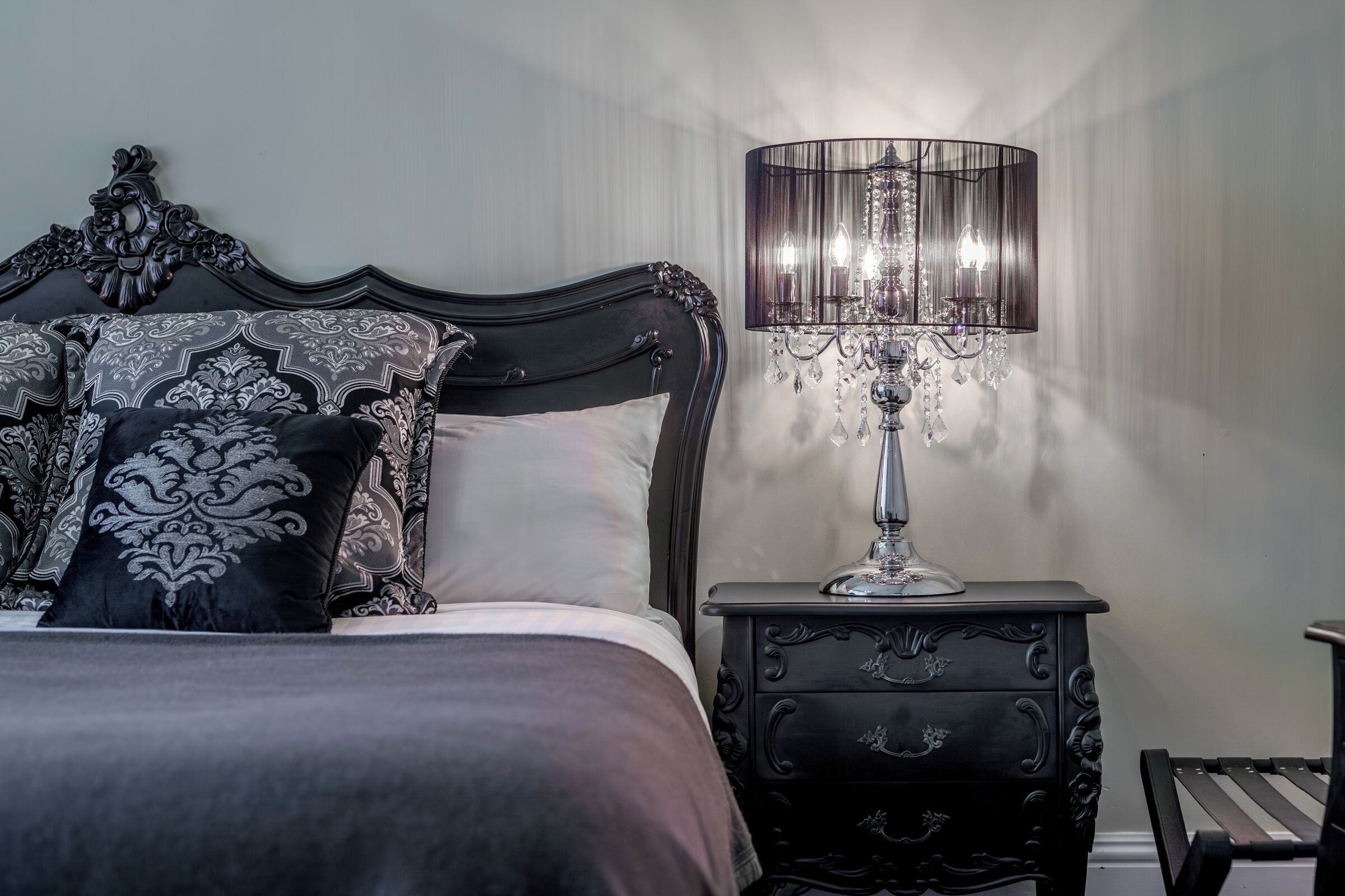Een slaapkamer met een hoofdeinde van donker hout, decoratieve kussens, een nachtkastje met een lamp en een bankje.