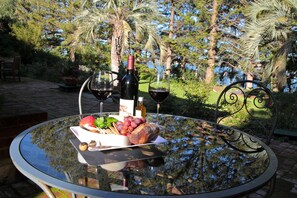 A glass table with a bottle of wine, two glasses, a bowl of fruit, and a loaf of bread in a garden setting.