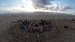 Un grupo de personas sentadas en círculo sobre una playa de arena, con el océano al fondo.