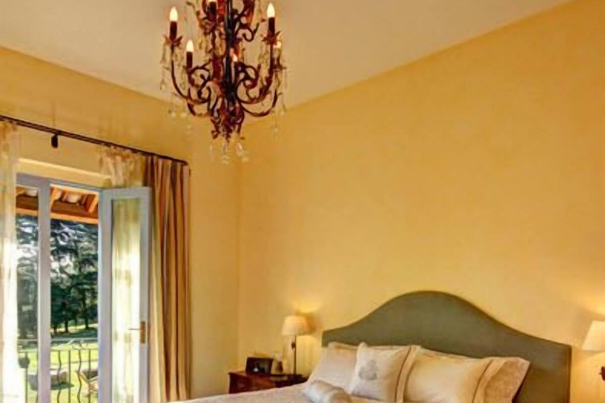 A bedroom with a large bed, a chandelier, a nightstand, and a view of a balcony.