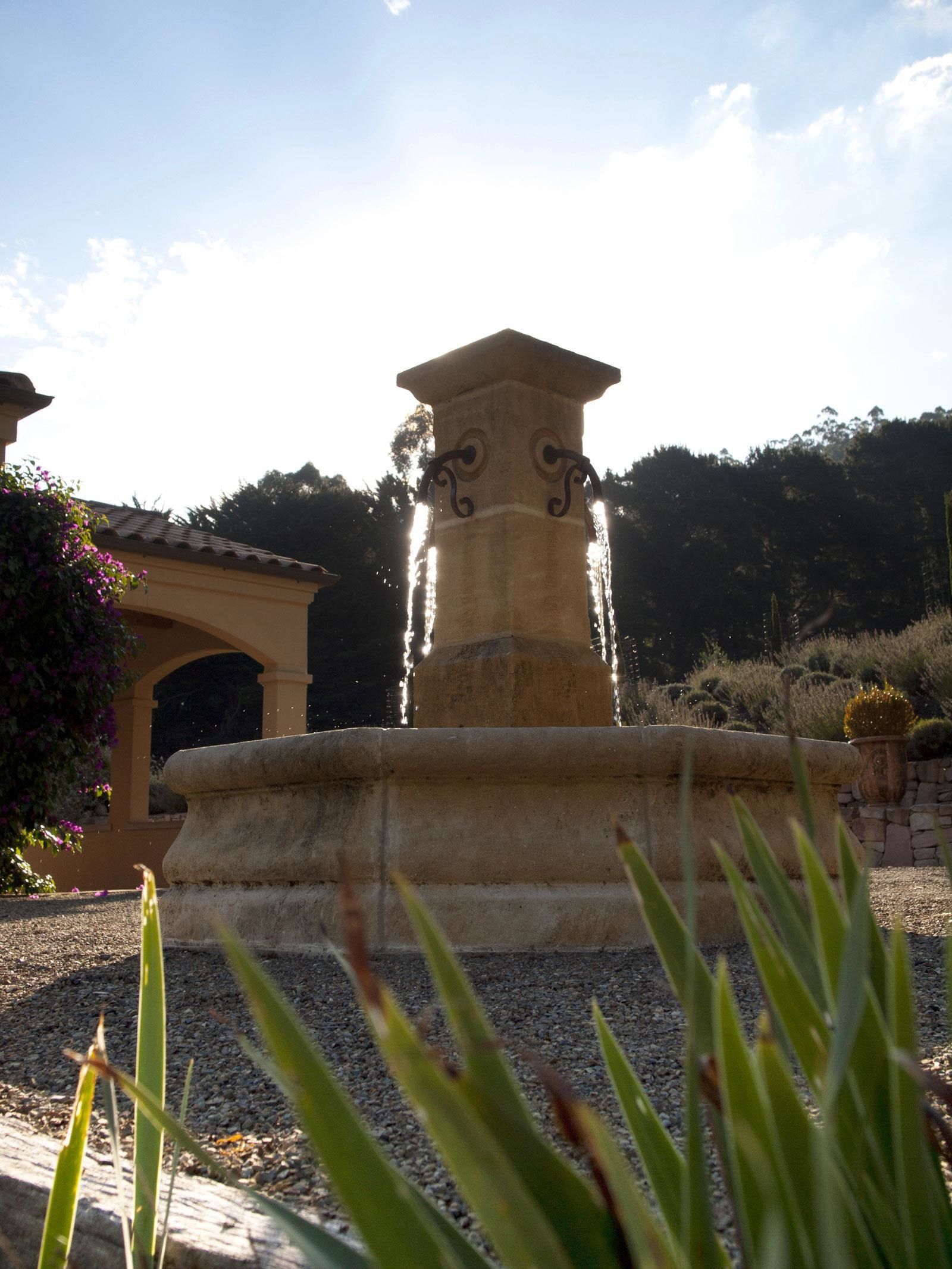A stone fountain with water spouting from the top, surrounded by gravel and plants.