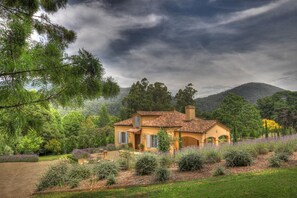 Uma casa com telhado de azulejos, rodeada por um jardim com diversas plantas e uma montanha ao fundo.