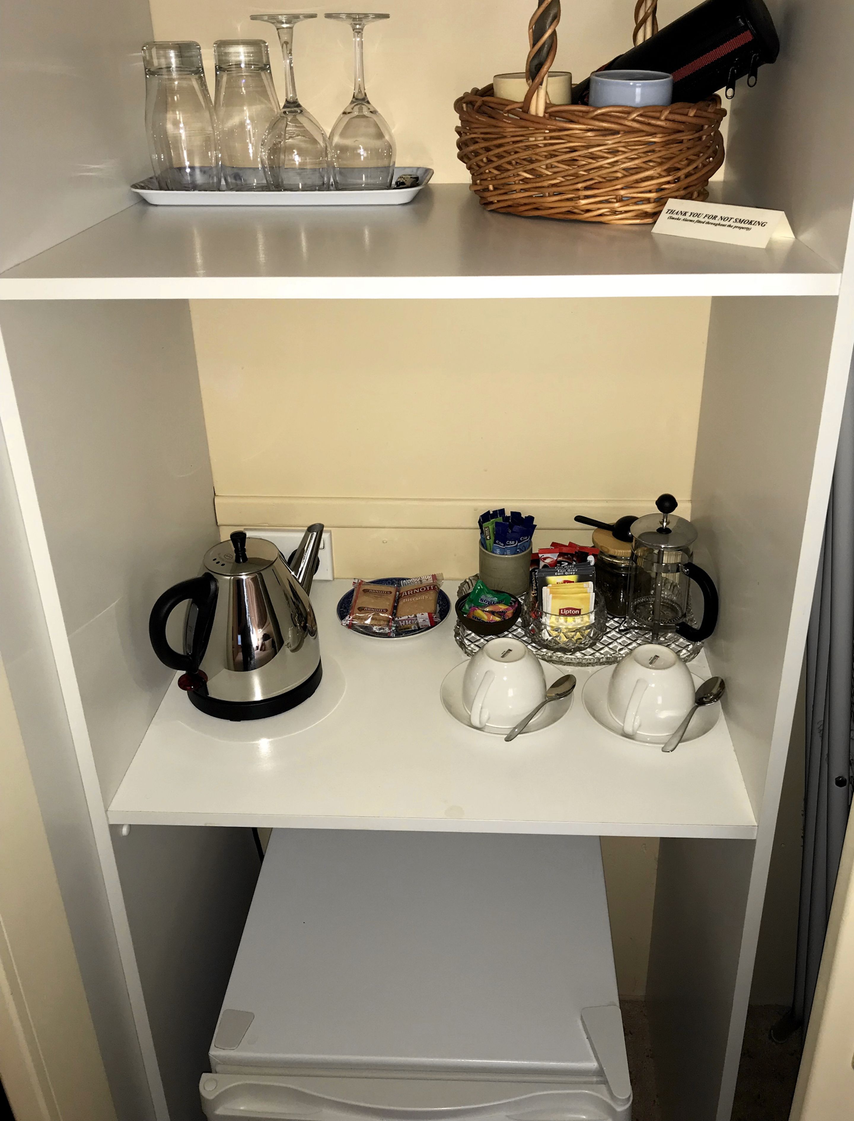 A kitchen area with a kettle, teapots, cups, and a basket containing a candle and a mug.