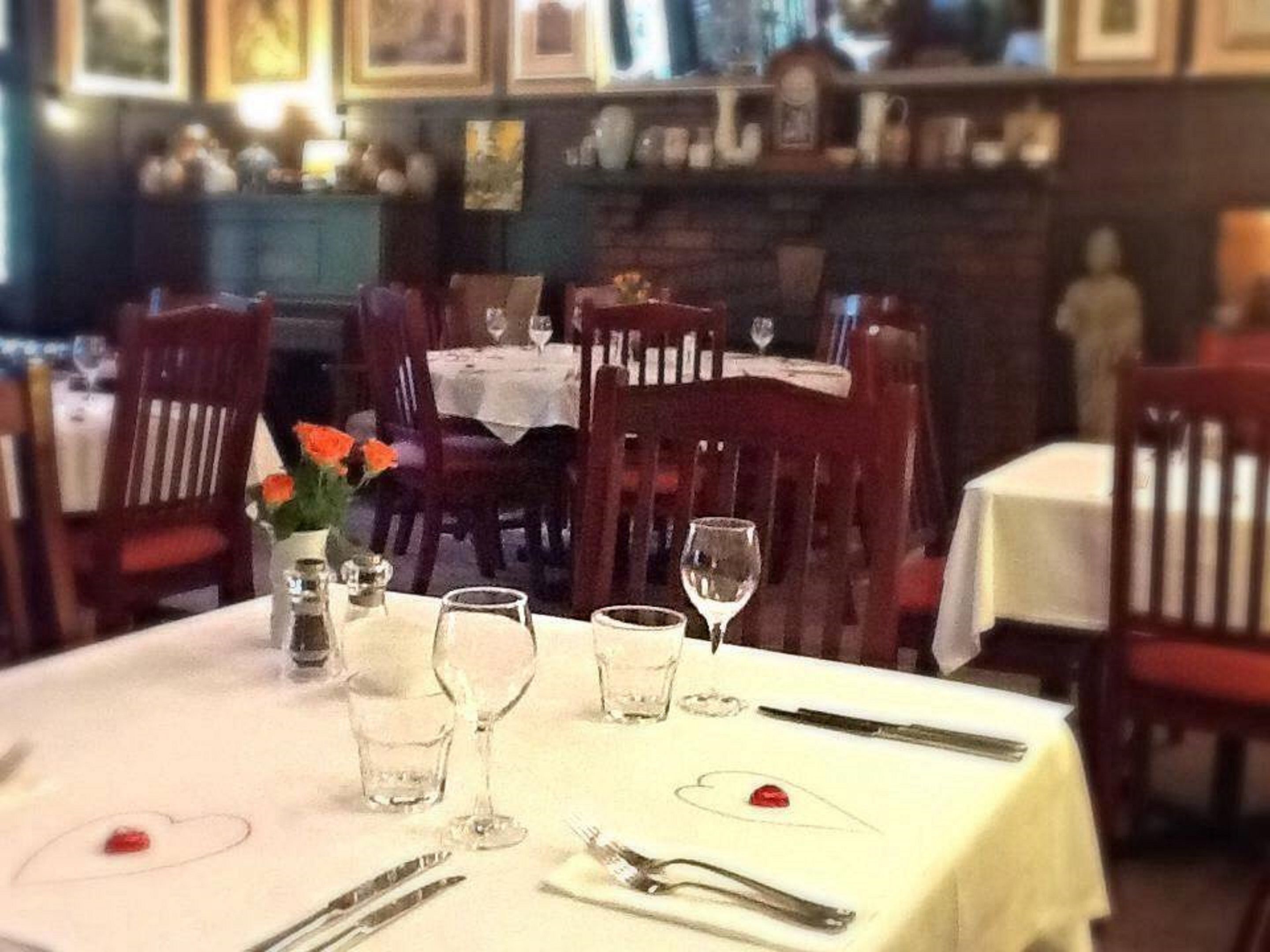 Un comedor con una mesa lista para dos personas, con mantel blanco, copas de vino, cubiertos y un florero con flores.