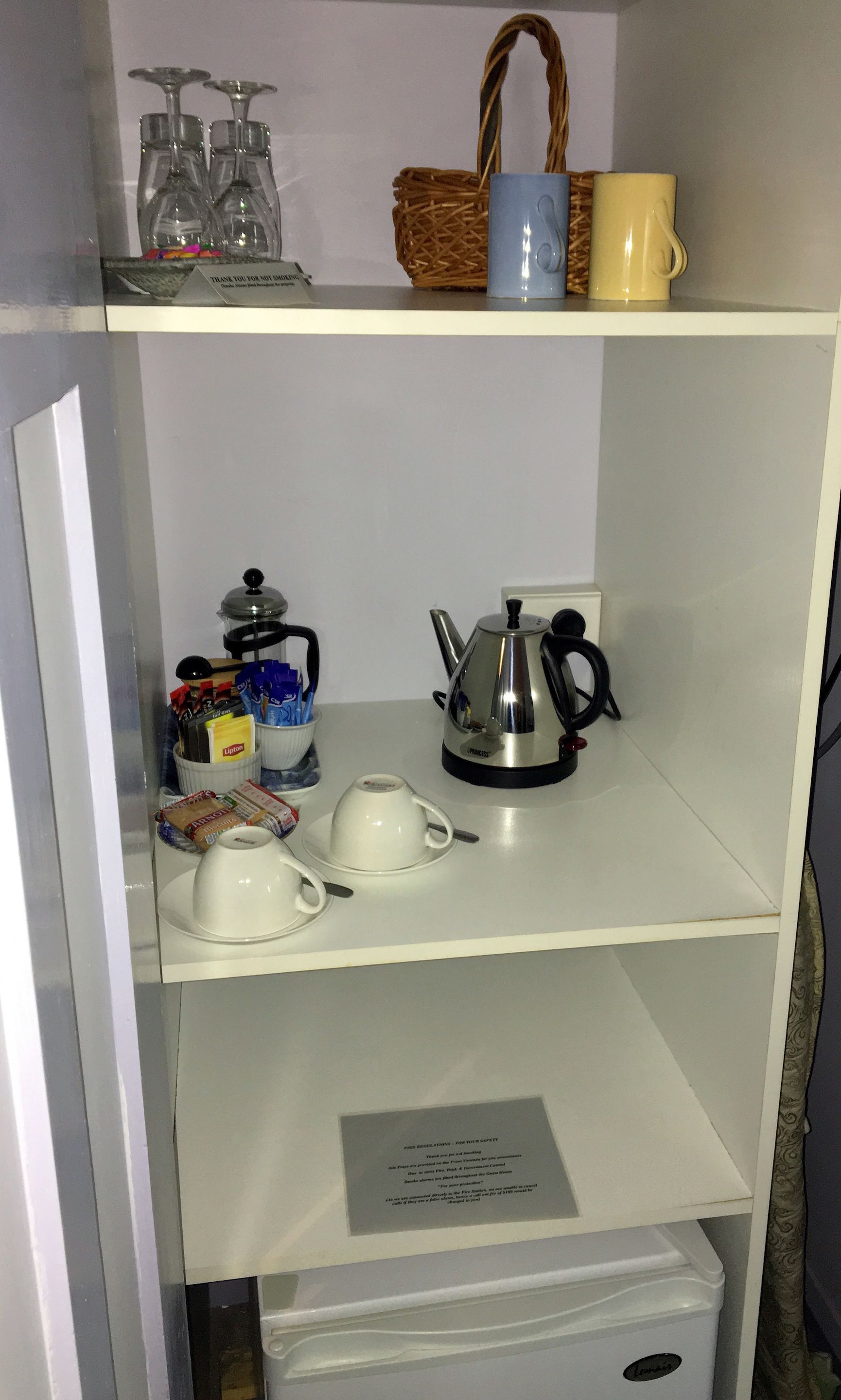 A kitchenette with a kettle, cups, and a basket on the shelf.