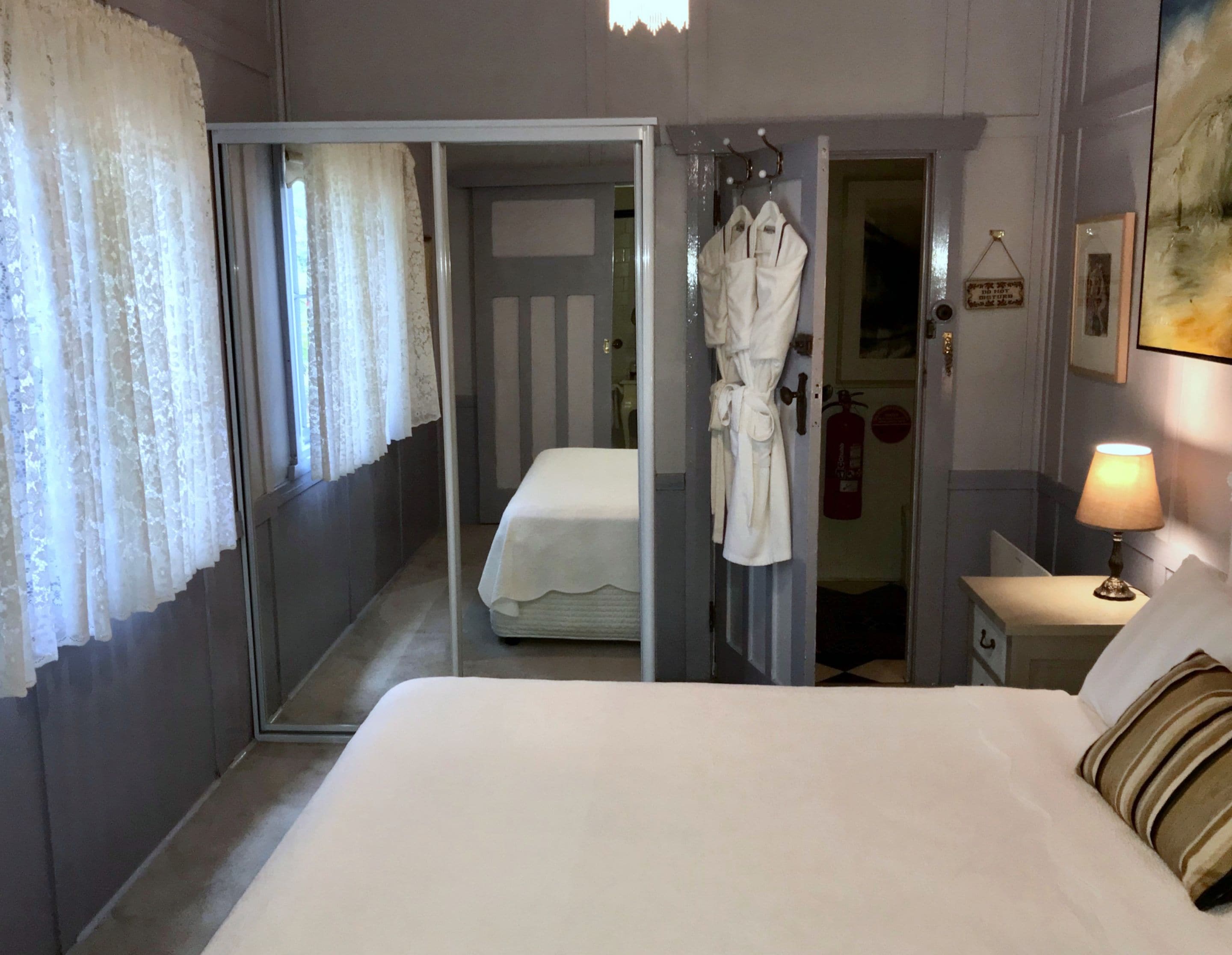 Een hotelkamer met een groot bed, een badjas hangend aan de deur, een bedlamp en een brandblusser aan de muur.