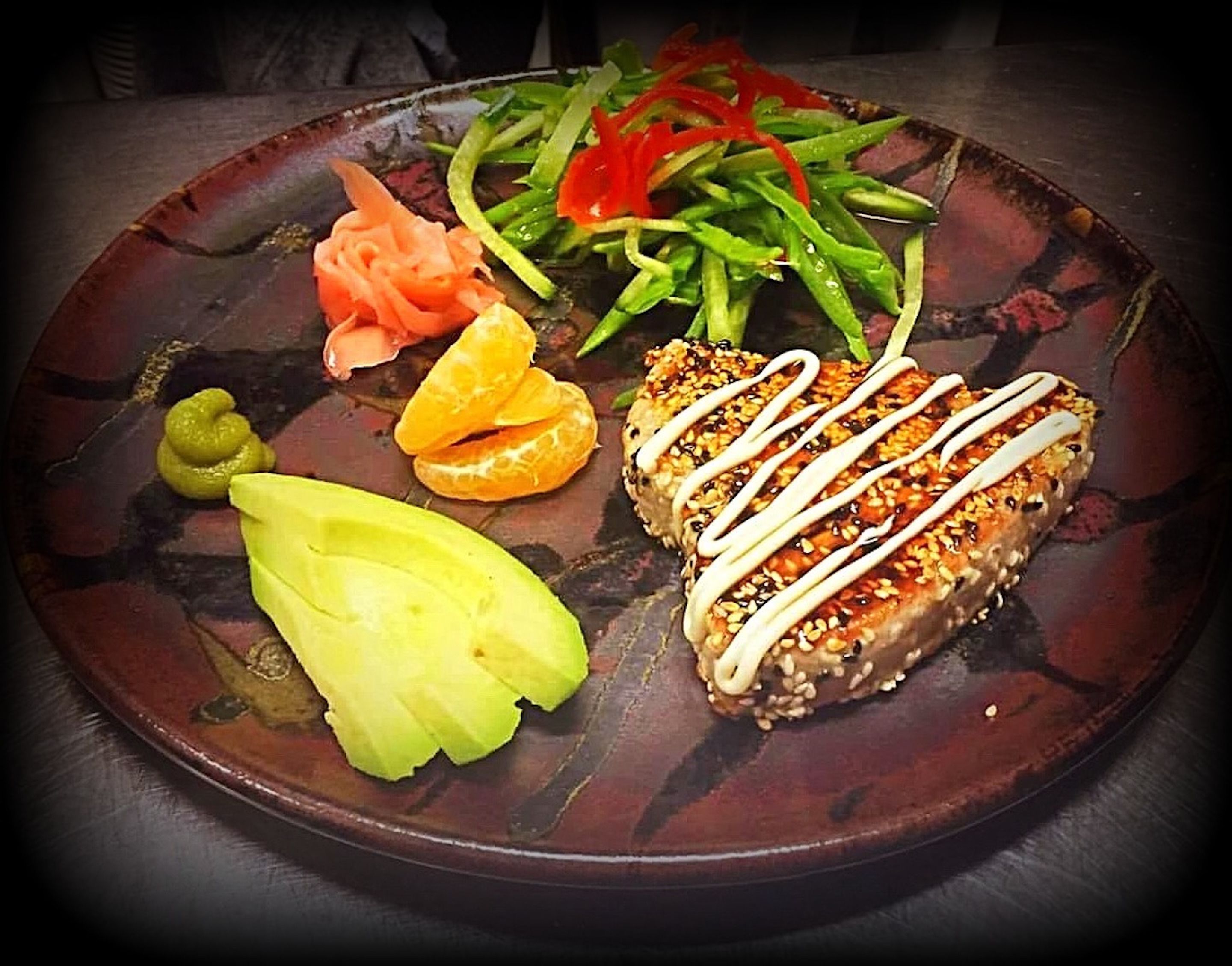 Un plato con diferentes alimentos, incluyendo una rebanada de pan con semillas de sésamo y salsa blanca, un vegetal verde, un vegetal amarillo y unas rodajas de naranja.