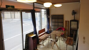 A room with a wooden table and chairs, a bench, a small cabinet, and a fan. There is a window with a view of the street outside. - Fuji Hostel (Onomichi)