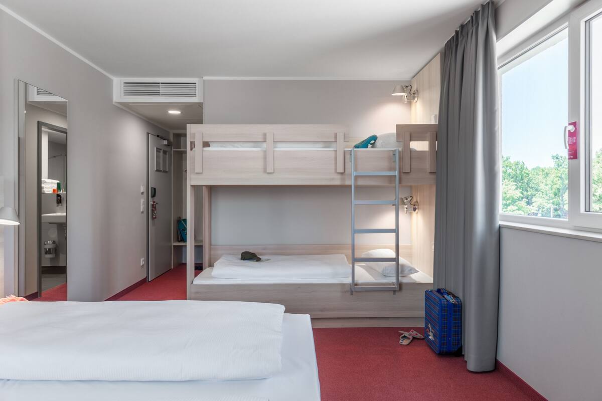 A hotel room with bunk beds, a red carpet, a window with curtains, and a blue suitcase.