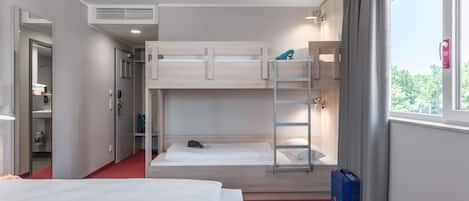 A hotel room with bunk beds, a red carpet, a window with curtains, and a blue suitcase.