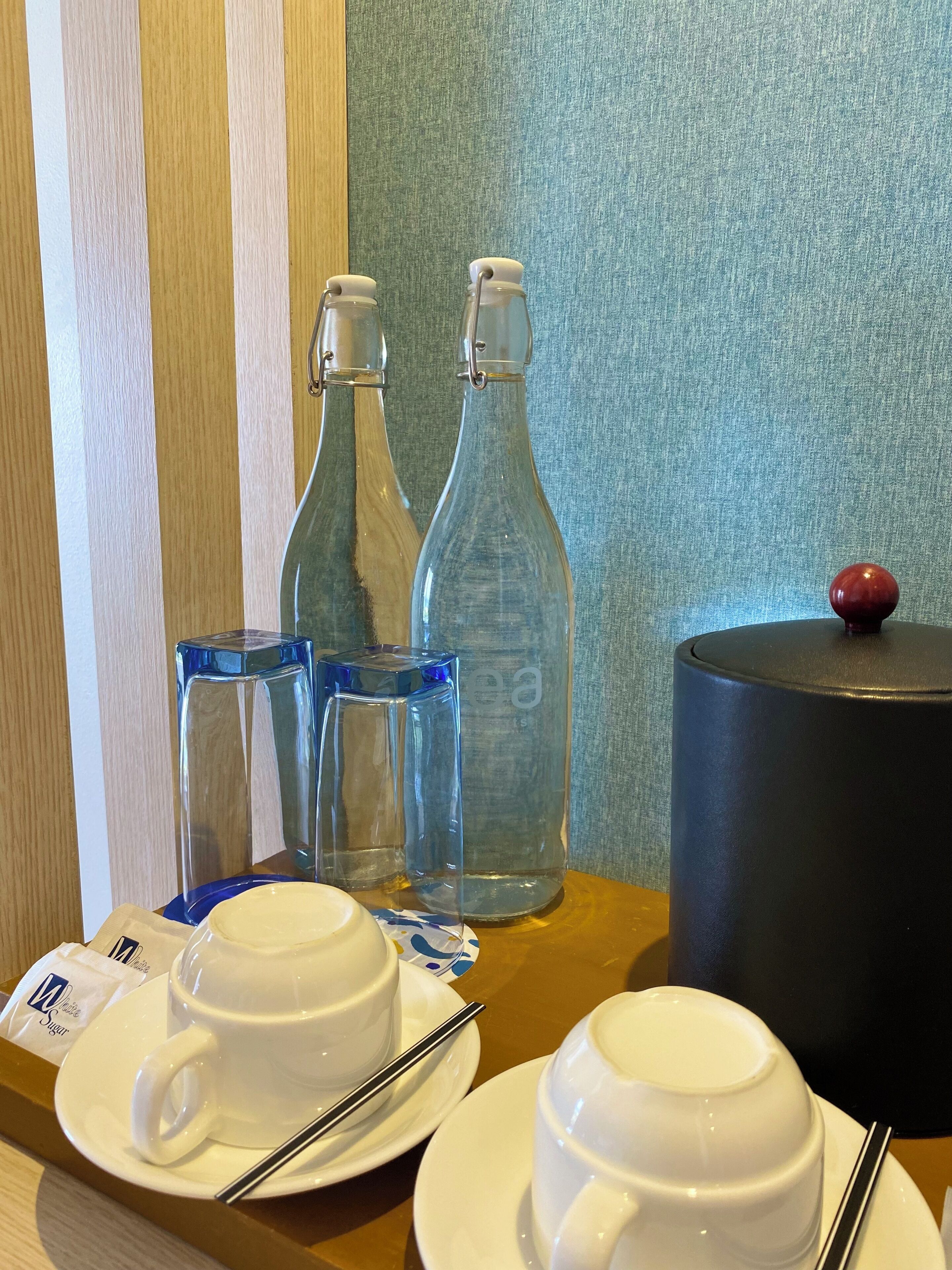 Una mesa con dos tazas blancas, un vaso azul y dos botellas transparentes con tapas blancas.