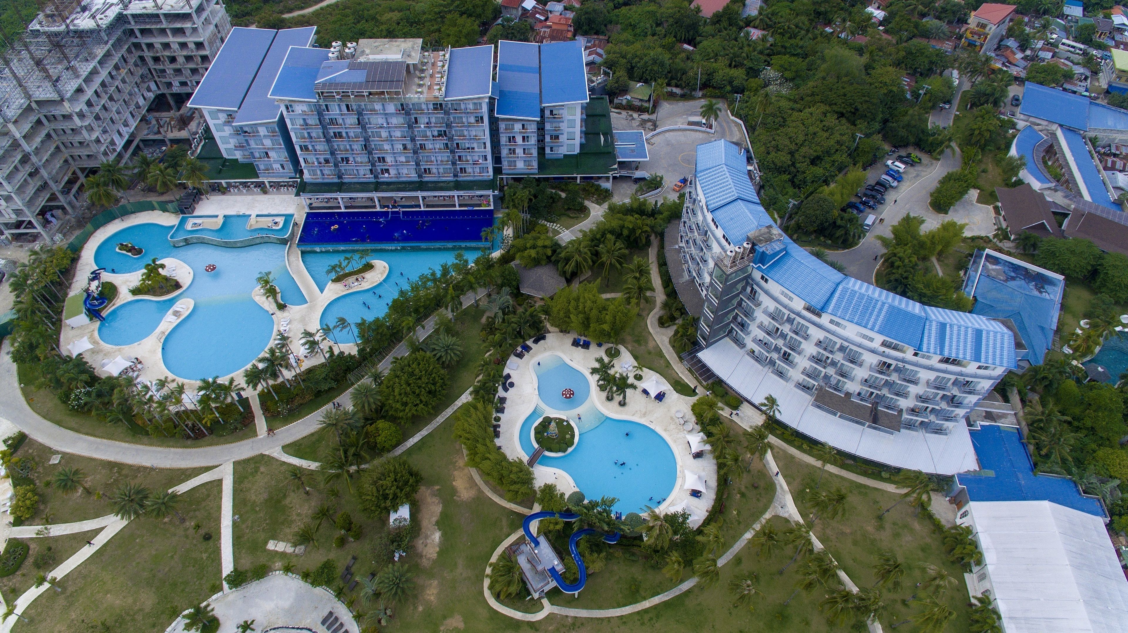 Vue aérienne d’un hôtel doté de plusieurs piscines, d’un grand bâtiment à toit incurvé et d’une végétation luxuriante environnante.