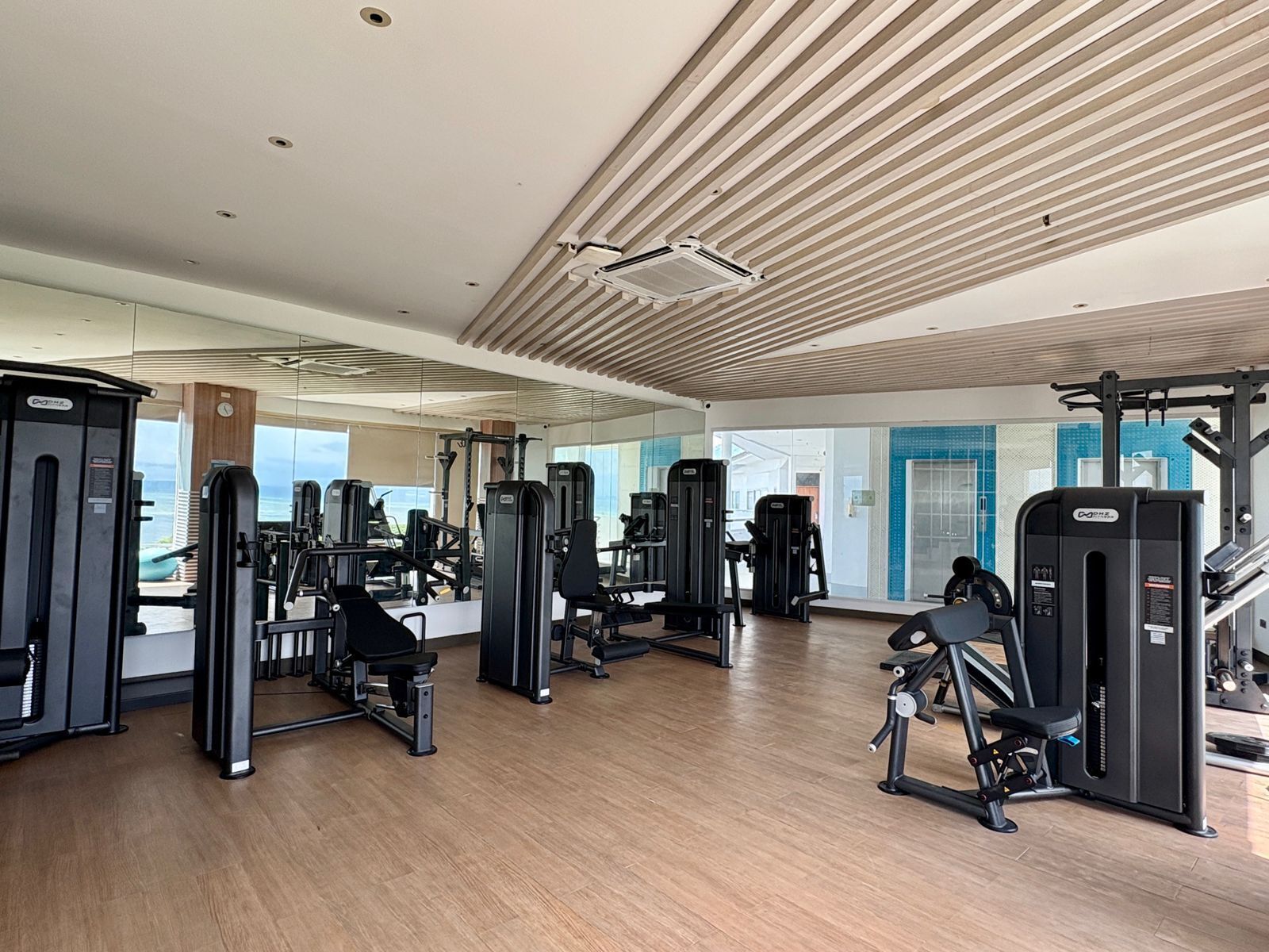 Un gimnasio con diversas máquinas de ejercicio, piso de madera y ventanas amplias.