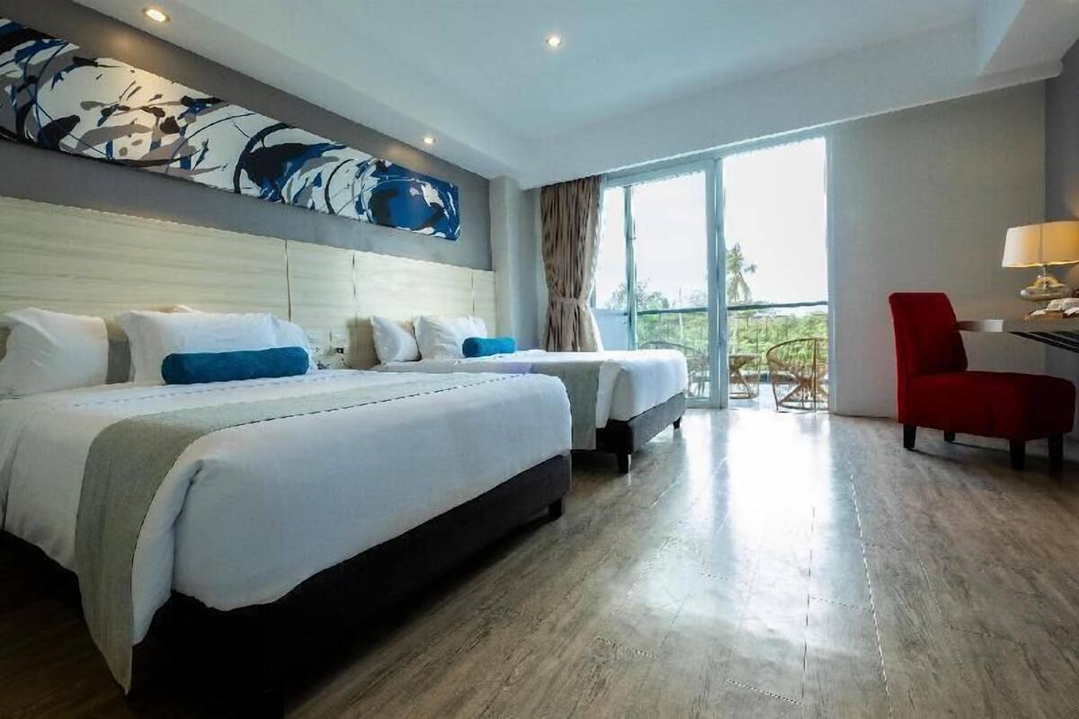 A hotel room with two beds, a red chair, a desk with a lamp, and a large window with a view.