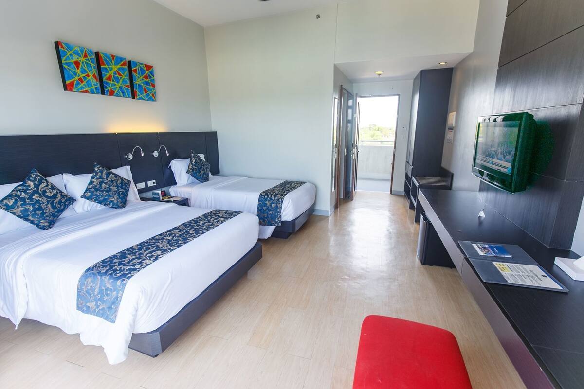 A hotel room with two beds, a red bench, a TV, and a view of the outdoors.