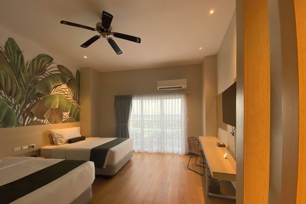 A hotel room with two beds, a ceiling fan, a TV, and a large window with curtains.
