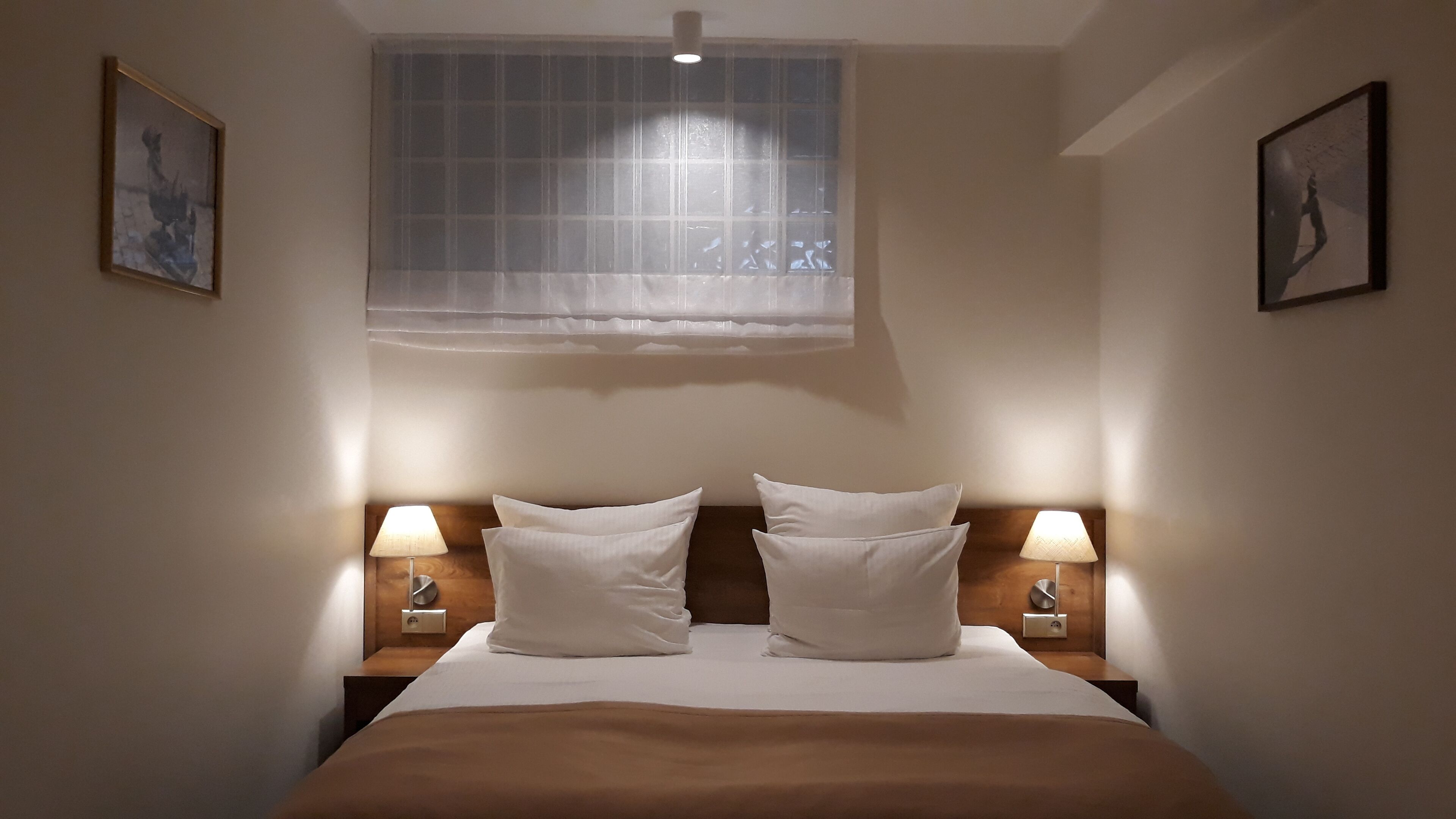 A hotel room with a bed, two bedside lamps, a window with a sheer curtain, and two framed pictures on the walls.