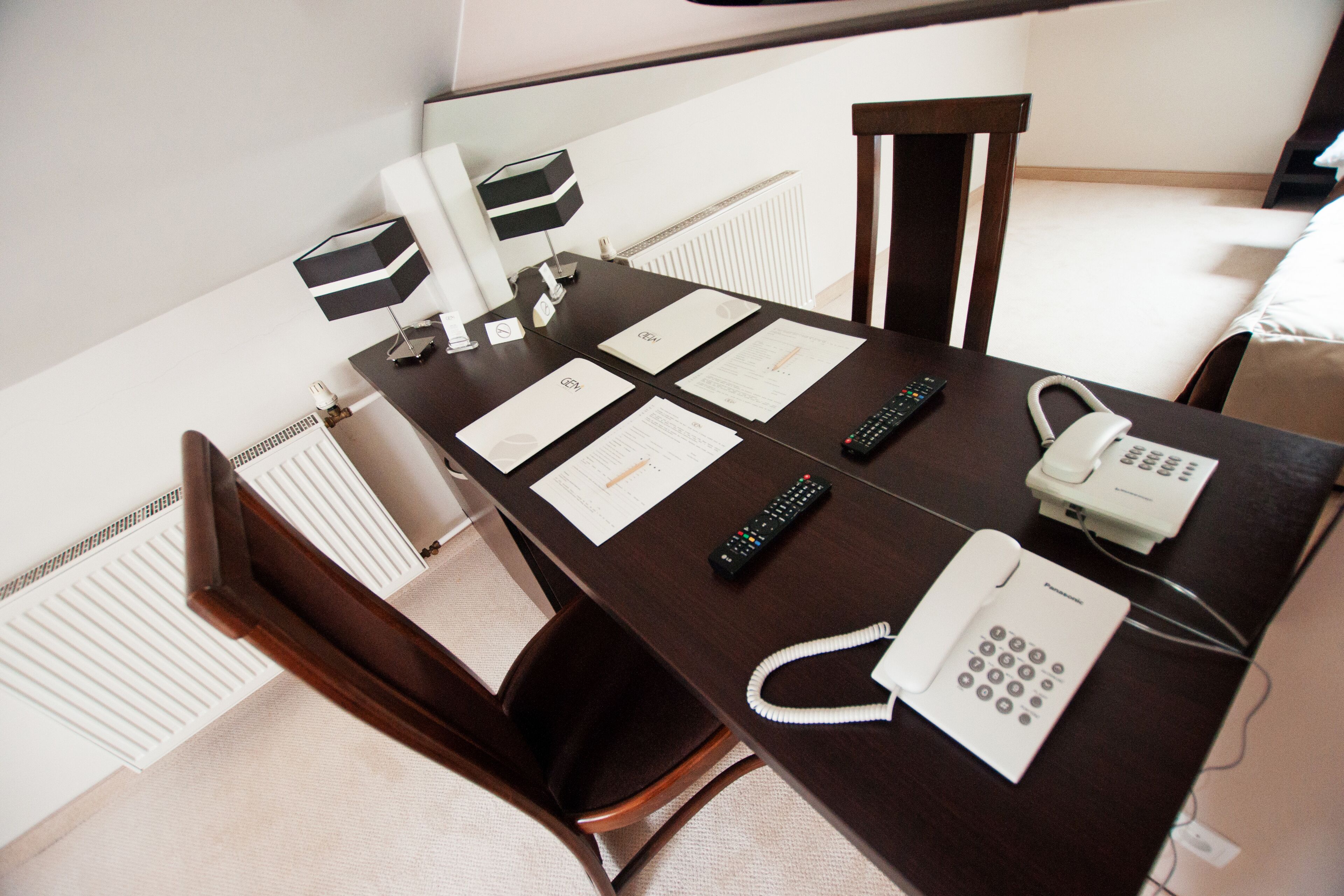 A hotel room with a wooden desk, a telephone, a remote control, and a lamp.