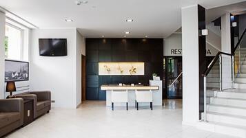 A modern hotel lobby with a reception desk, a flat-screen TV, a seating area with a sofa, and a staircase.