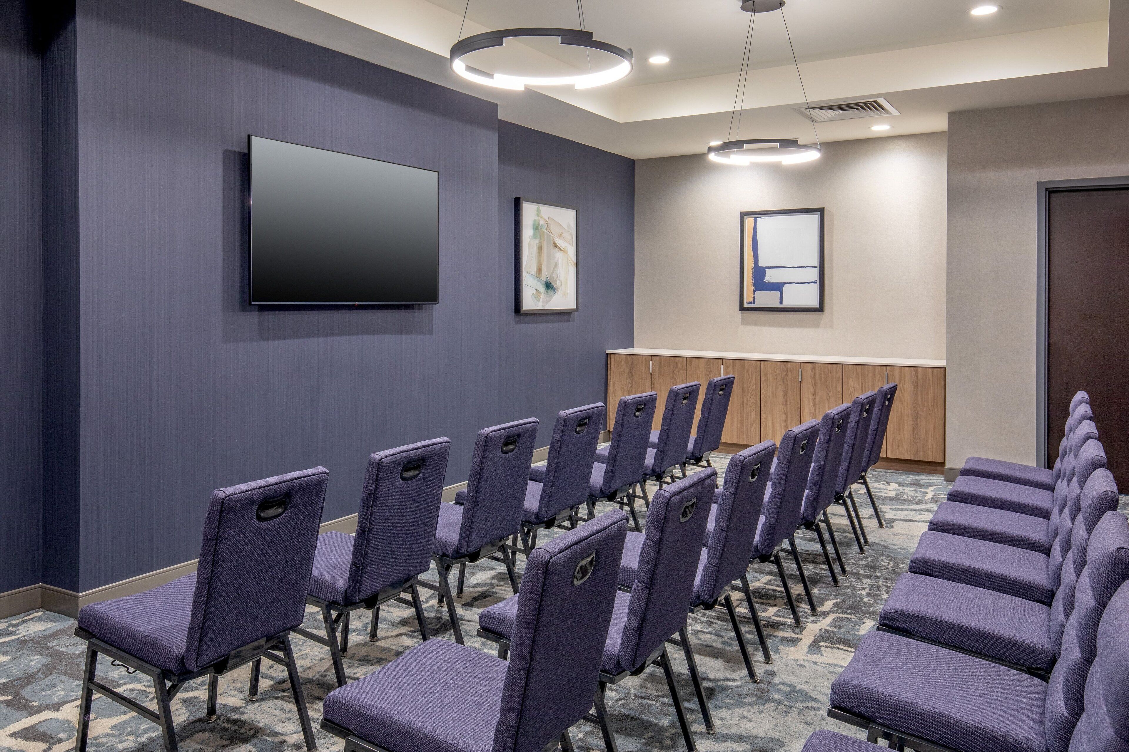 A conference room with rows of purple chairs, a flat-screen TV, and framed artwork on the walls.