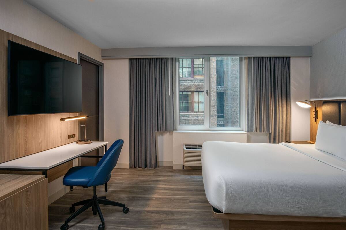 Hotelkamer met een bed, bureau, stoel, televisie en groot raam met gordijnen.