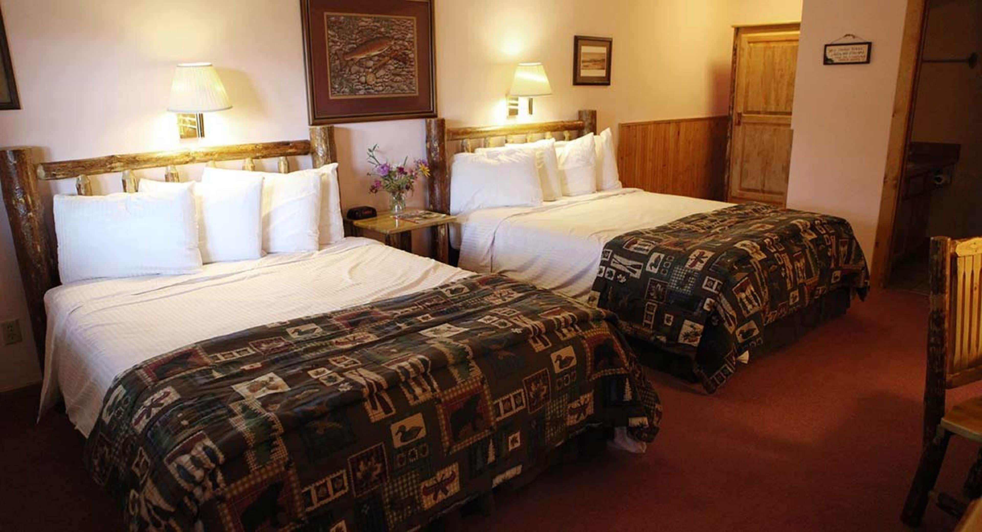 Ein Hotelzimmer mit zwei Betten, jeweils mit weißen Leintüchern und einer gemusterten Decke. Es gibt Nachttische mit Lampen, ein gerahmtes Bild an der Wand und einen Holzschrank.