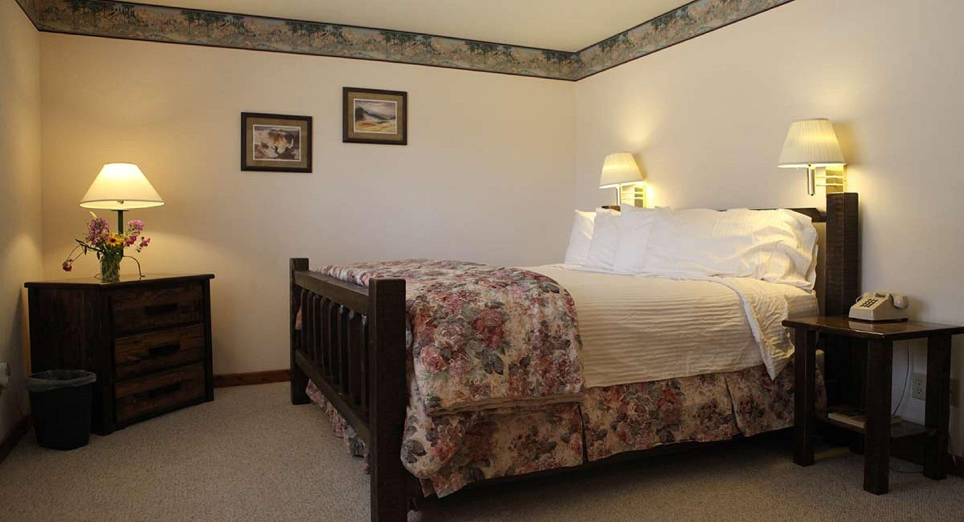 A bedroom with a wooden bed, floral bedspread, two bedside lamps, a nightstand with a lamp, and a trash can.