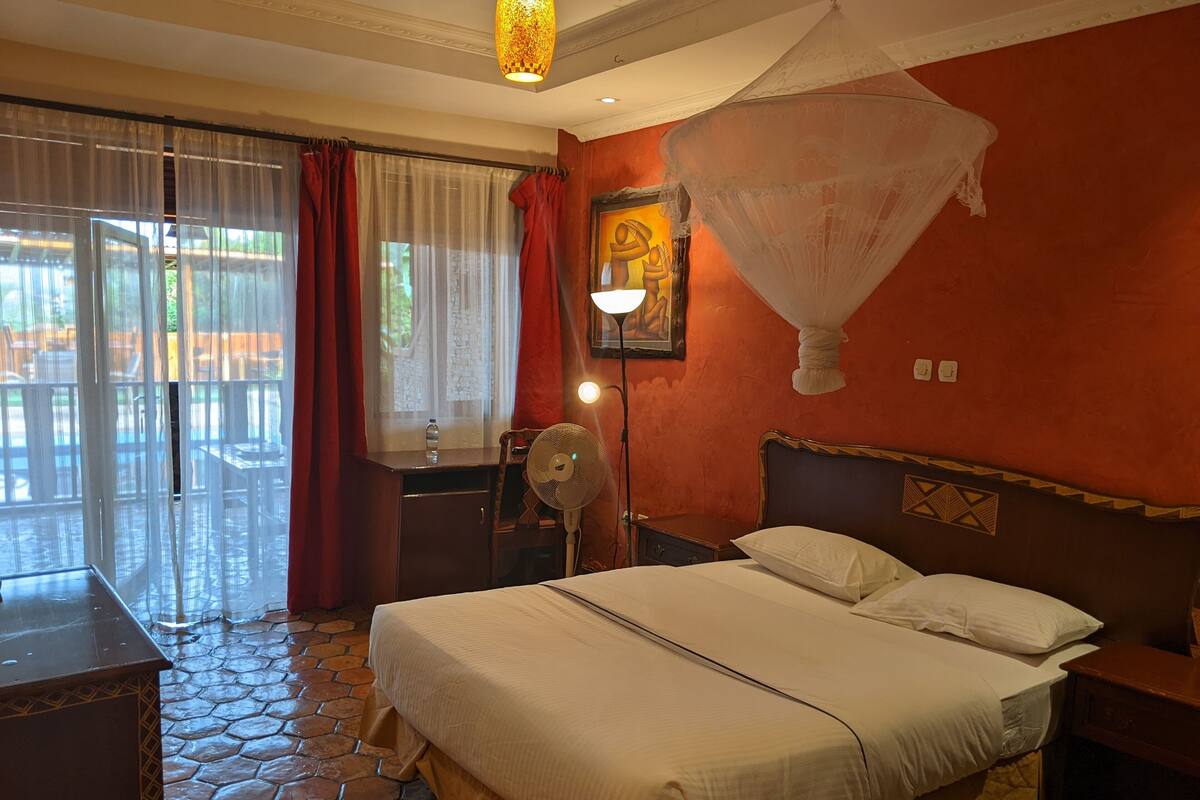 A bedroom with a bed, a nightstand, a bedside table, a fan, a window with curtains, and a painting on the wall.