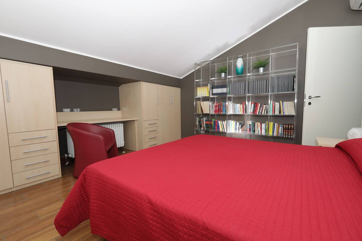A bedroom with a red bedspread, a bookshelf, a desk, and a wardrobe.