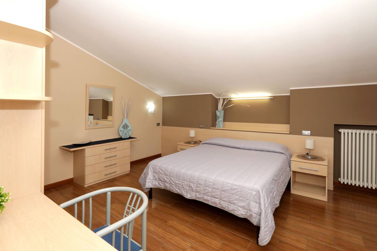 A bedroom with a bed, bedside tables, a chair, a dresser, a mirror, and a radiator.