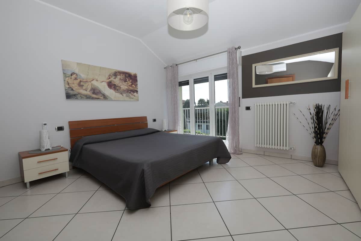 A bedroom with a large bed, a nightstand, a painting on the wall, a radiator, and a window with curtains.