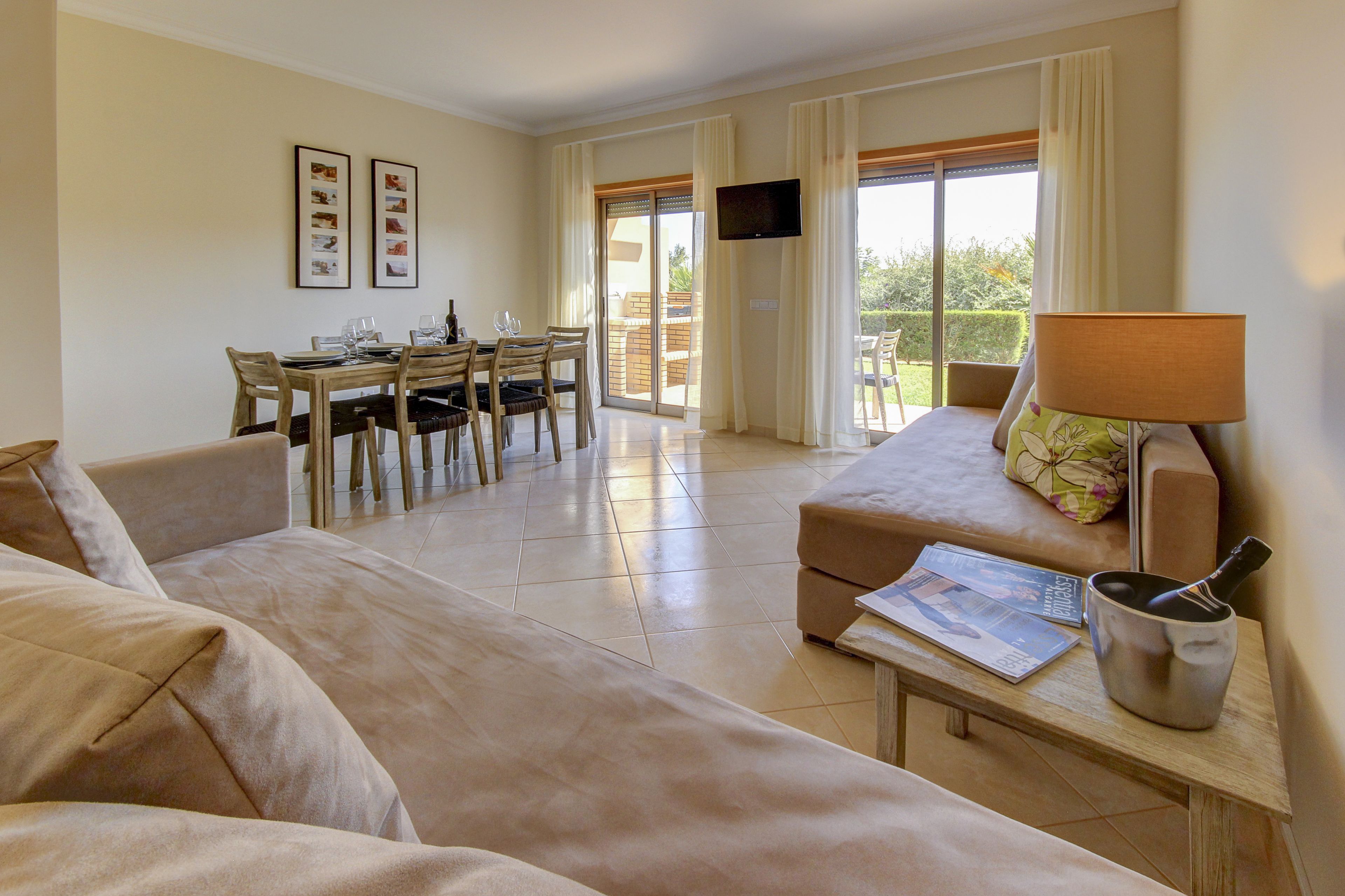 A spacious living area with a sofa, a dining table with chairs, a flat-screen TV mounted on the wall, and a balcony with a view.