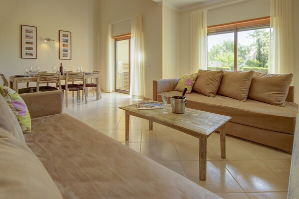 A spacious living room with a wooden coffee table, a sofa, and a dining area with chairs and a table. - Pinheiros da Balaia (Albufeira)