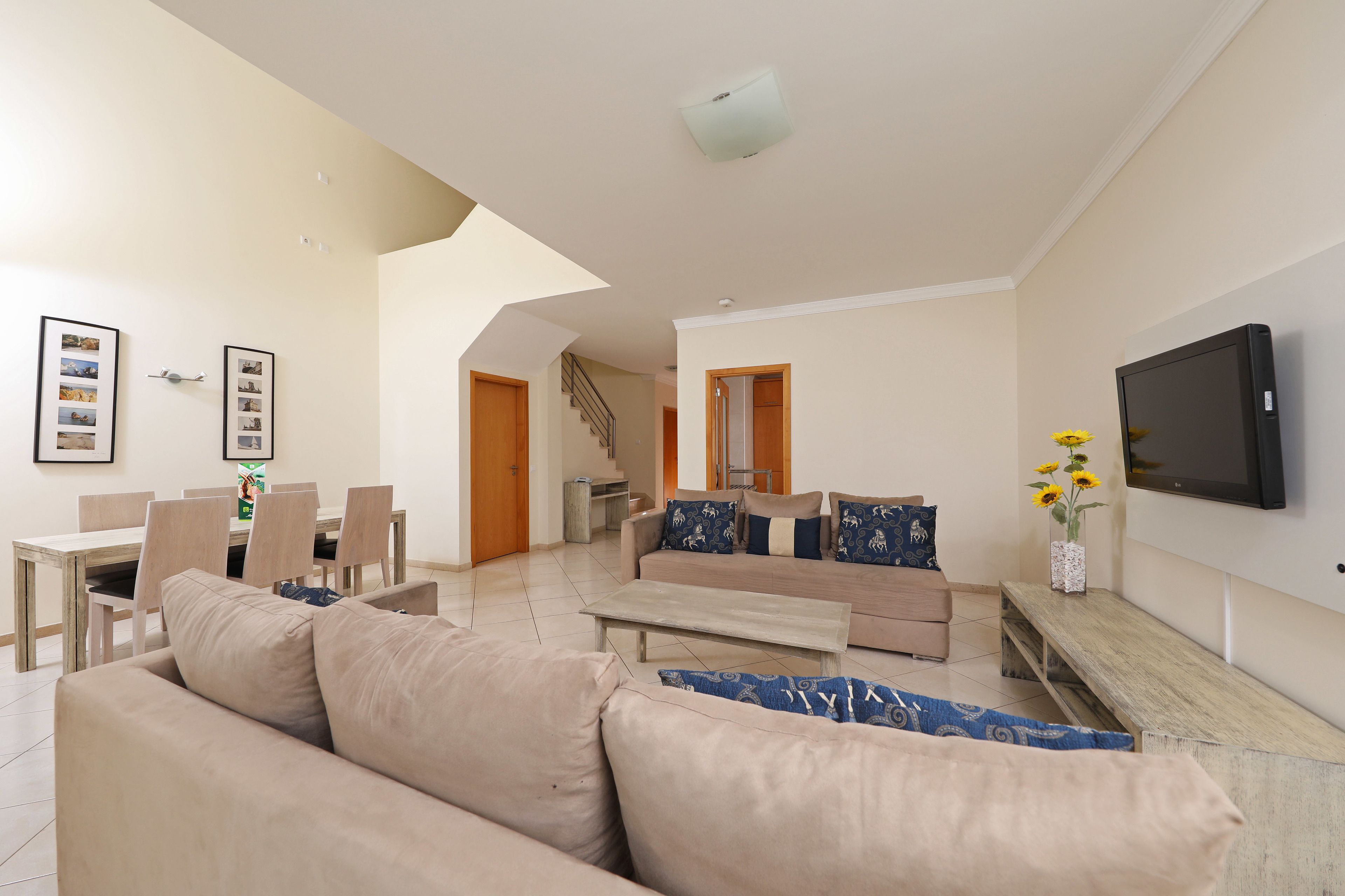 A spacious living room with a sofa, coffee table, and dining area. There is a staircase leading to another level, and a flat-screen TV mounted on the wall.