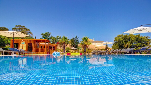 Piscina com chão azul em azulejos, espreguiçadeiras, guarda-sóis e um edifício de madeira com um letreiro.