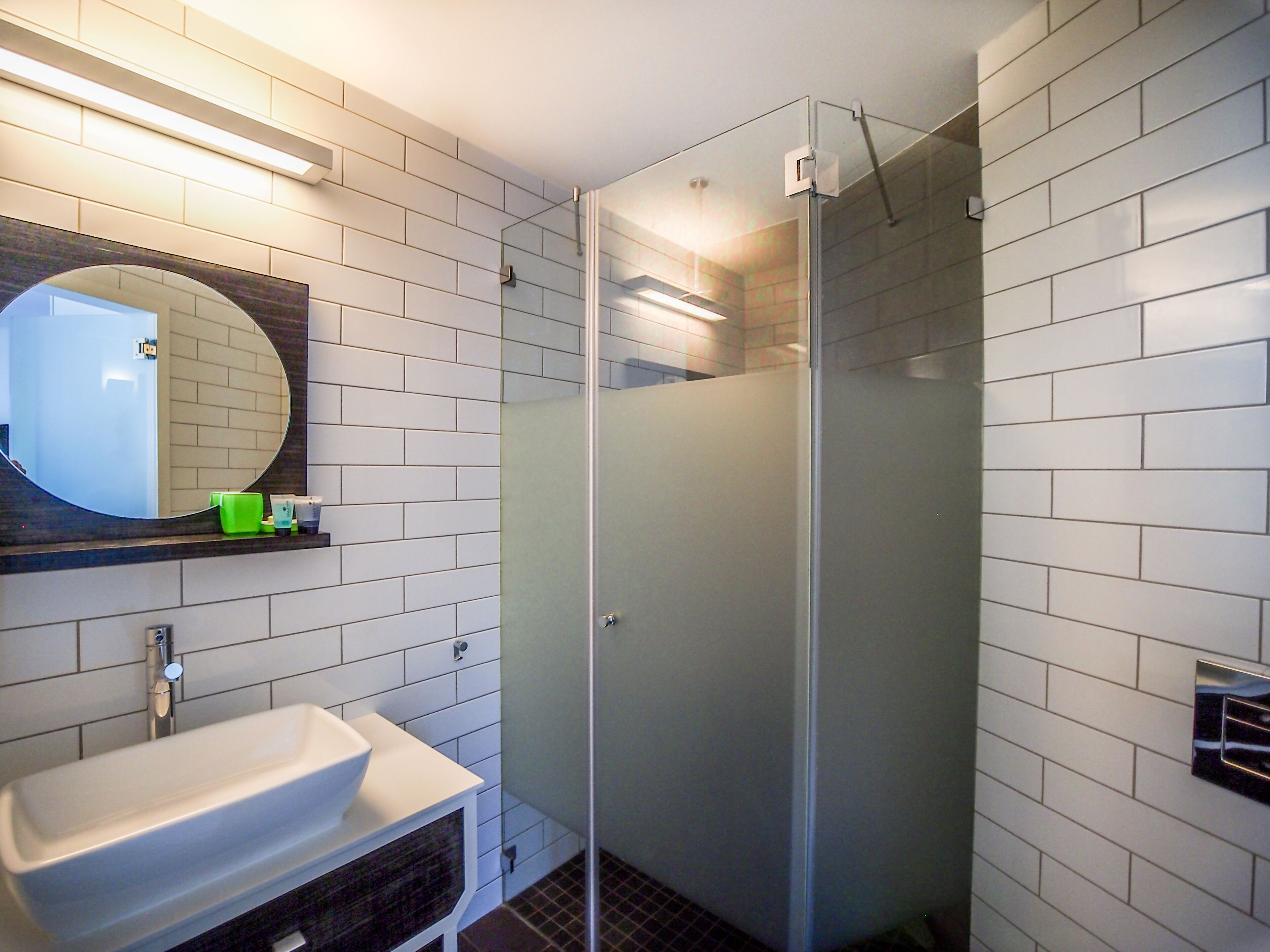 Une salle de bain moderne avec des carreaux de métro blancs, une douche en verre, un lavabo blanc et un miroir rond.