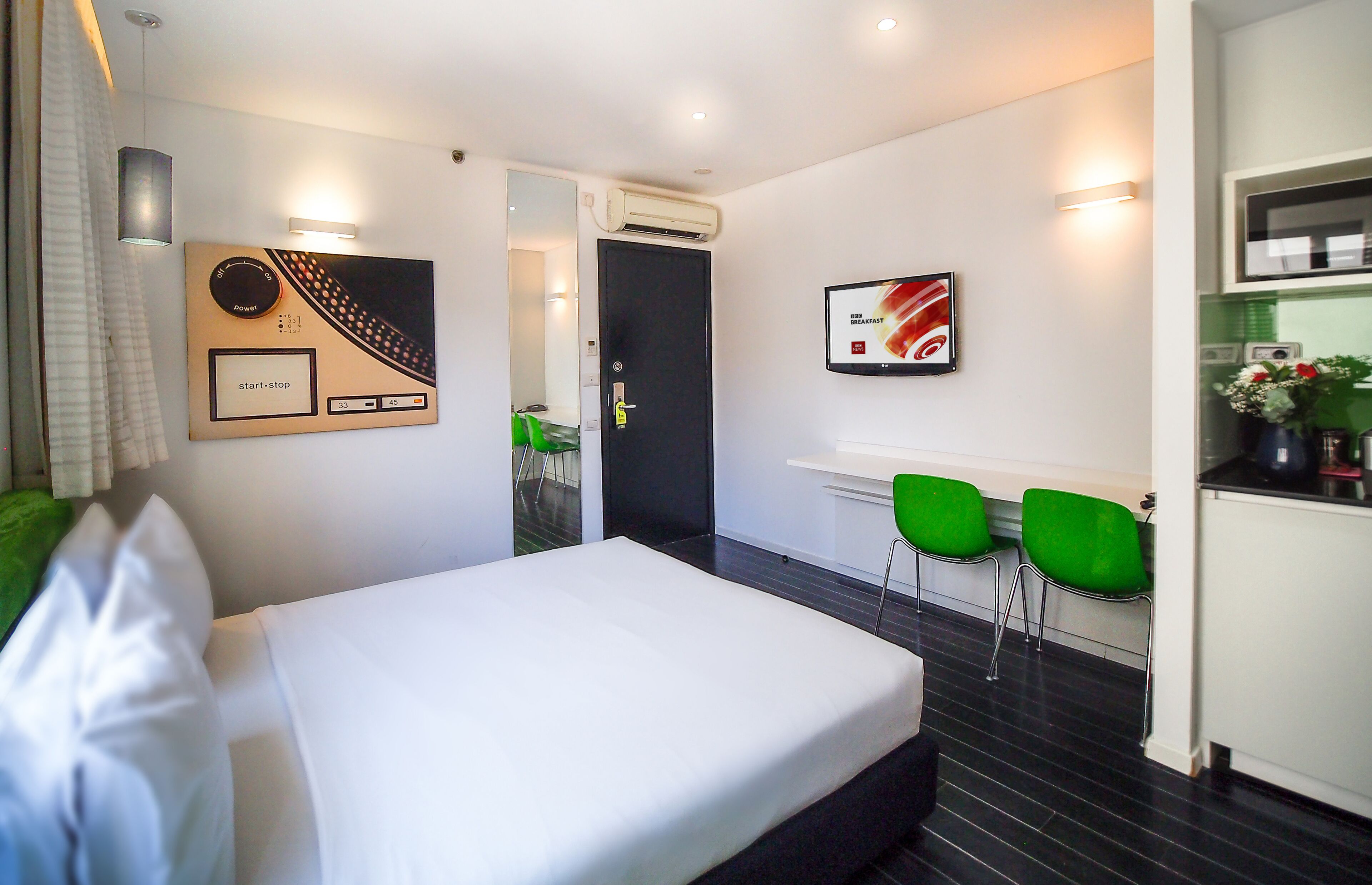 A hotel room with a bed, a desk, two green chairs, a wall clock, and a framed picture.