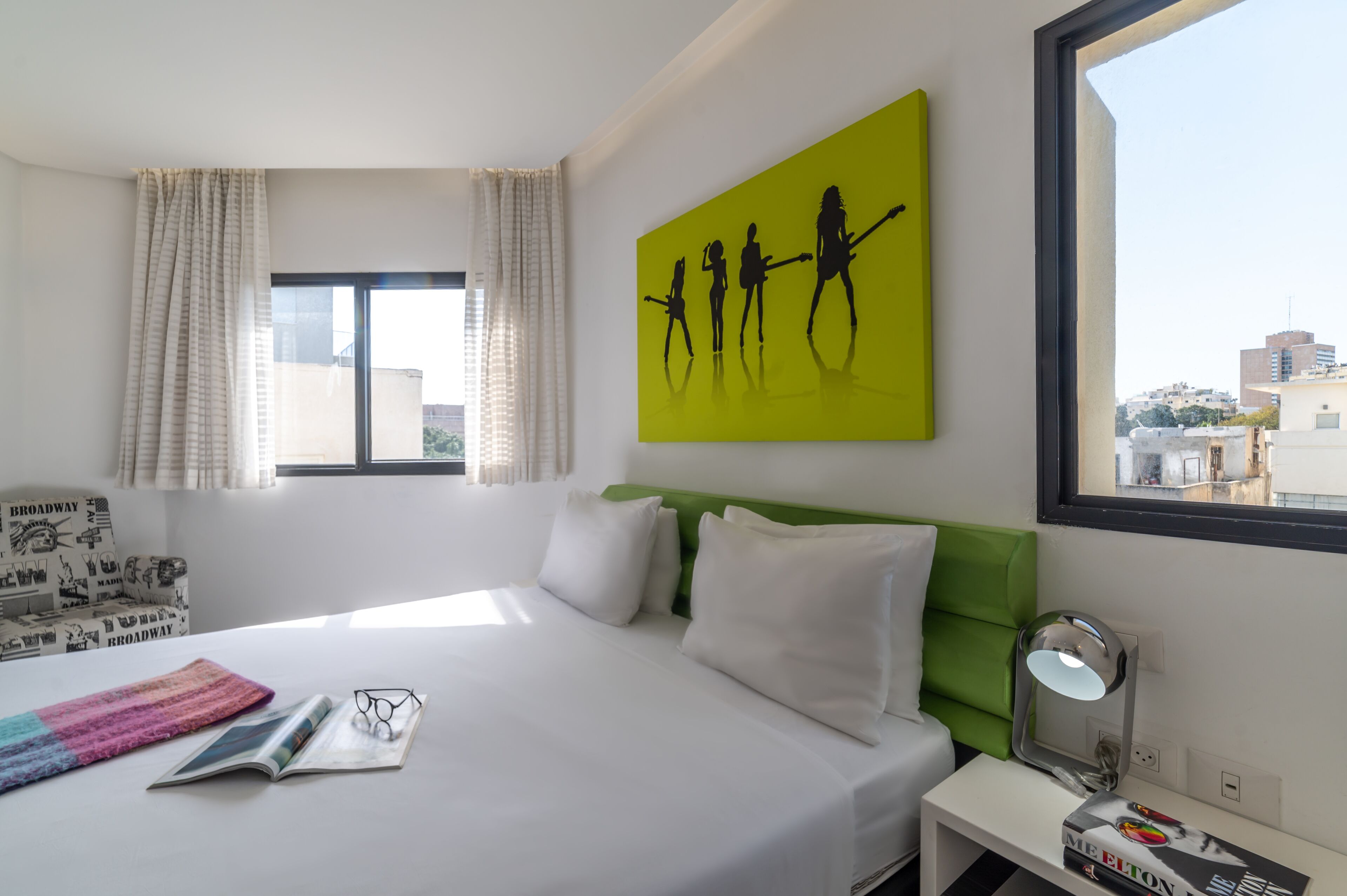 A hotel room with a bed, a green headboard, a window with curtains, a painting on the wall, a bedside table with a lamp, and a magazine on the bed.