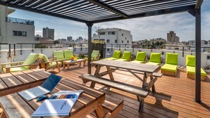 A rooftop terrace with wooden furniture, green cushions, and a table with a laptop and magazine. - 130 Rock Apartments (Tel Aviv)