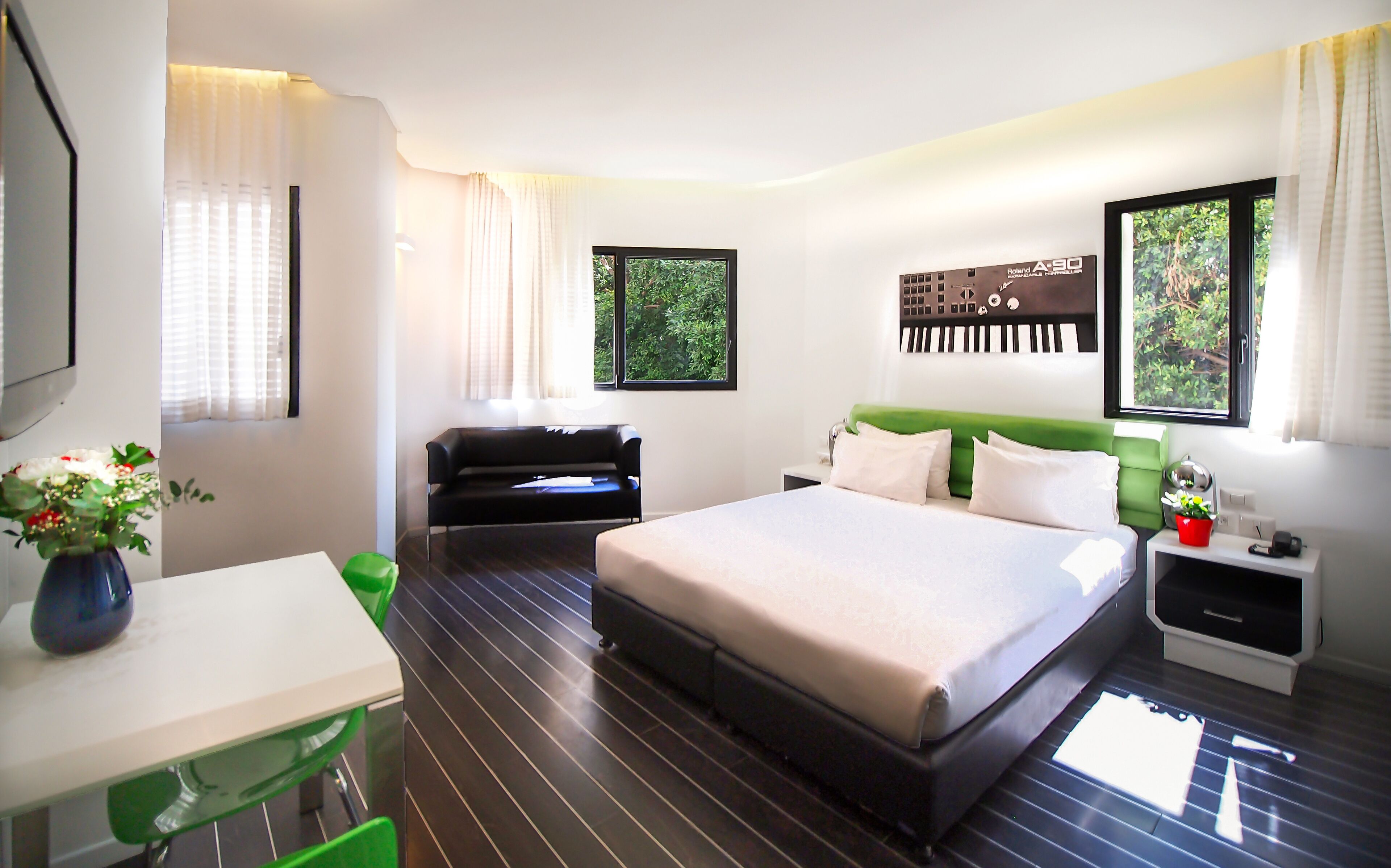 A modern hotel room with a bed, a desk, a chair, and a small table. There are windows with curtains, a green headboard, and a wall-mounted artwork.