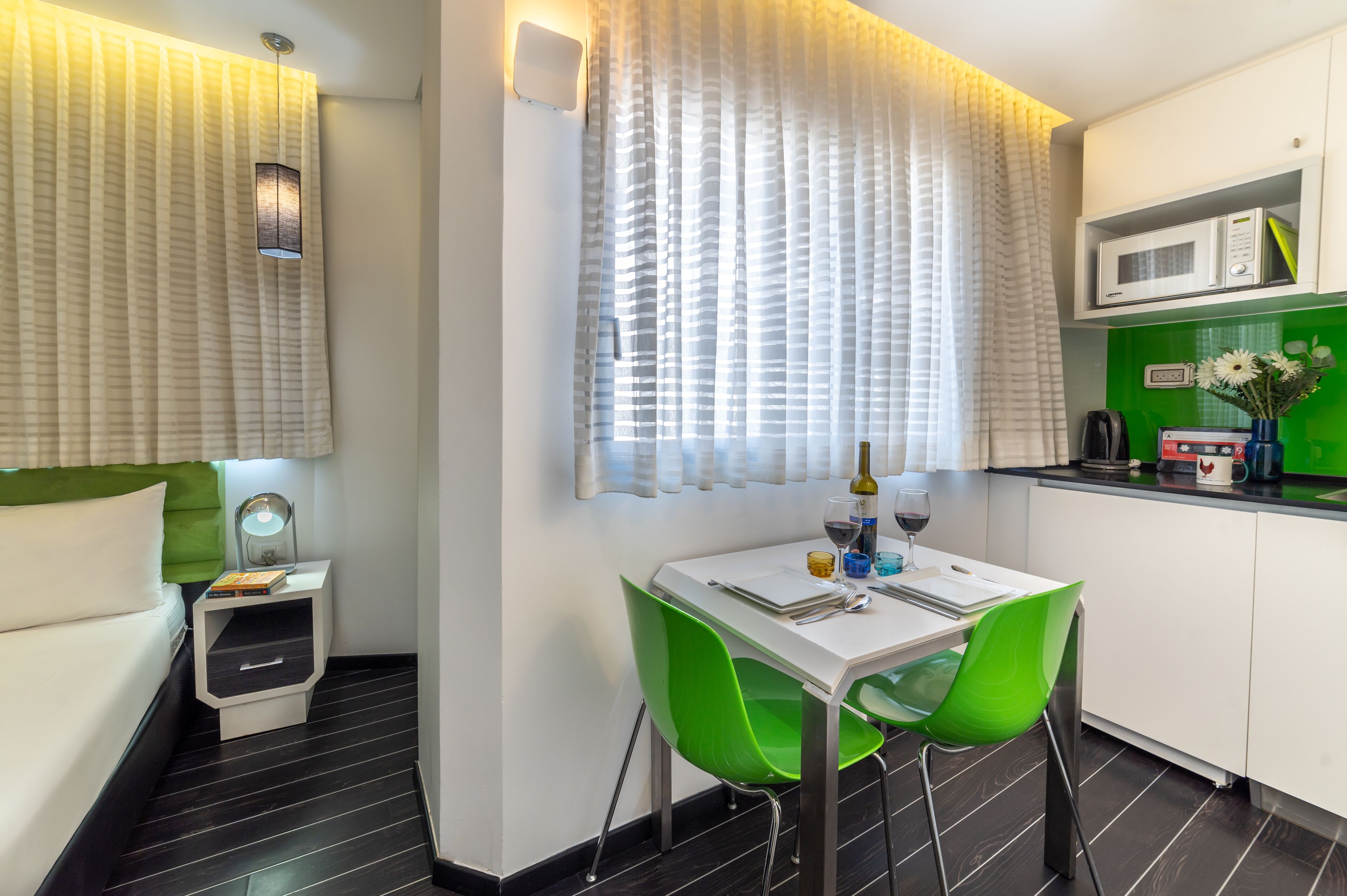 Une chambre d’hôtel moderne avec un lit, une table de chevet, des chaises vertes, une table à manger, un four à micro-ondes et un mur vert.