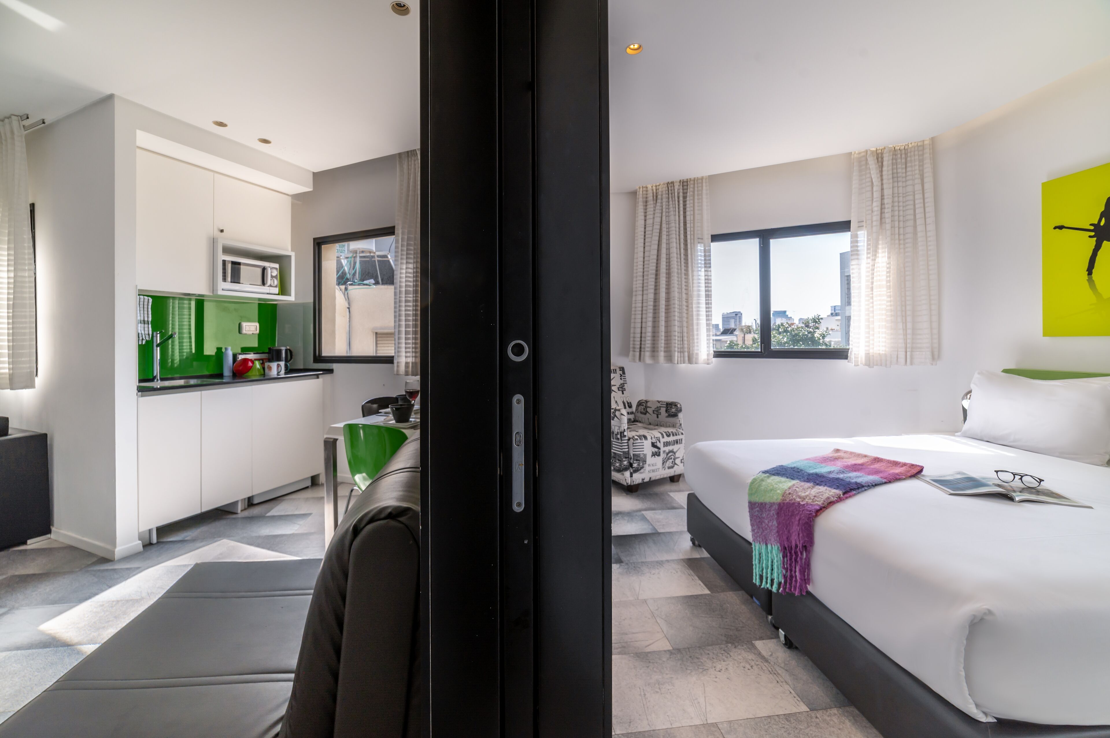 A modern hotel room with a bed, a small kitchenette, and a view of the city.