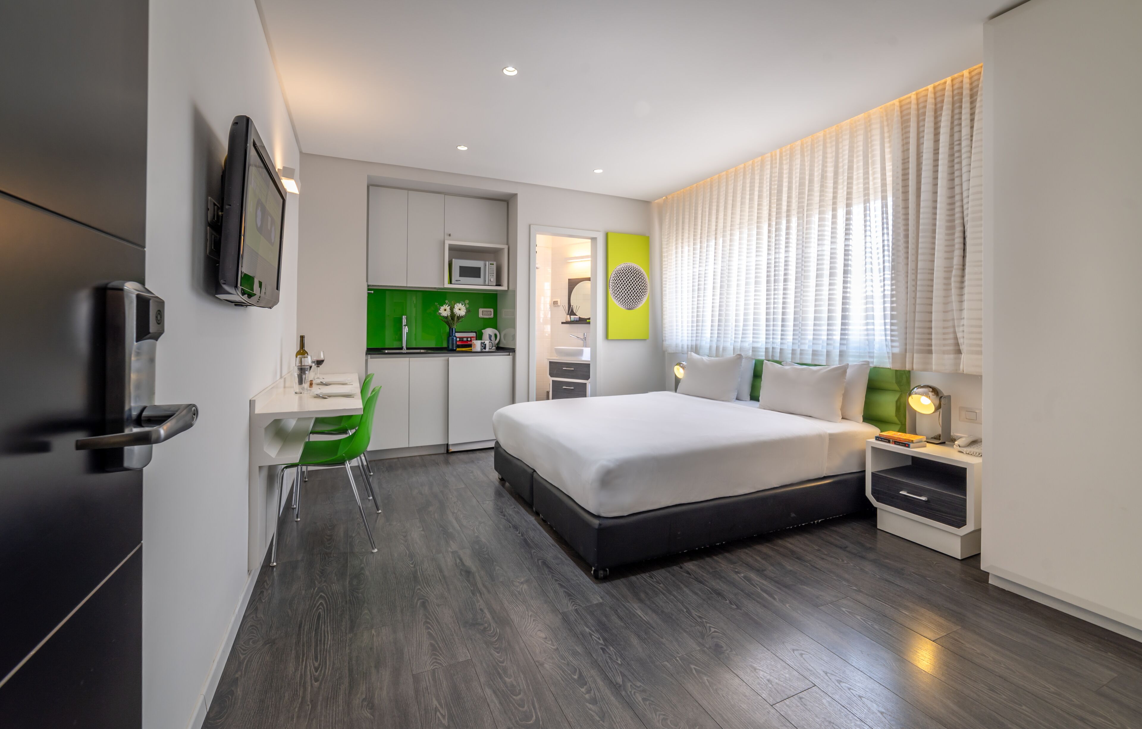 A modern hotel room with a large bed, a desk, a chair, a TV, and a kitchenette.