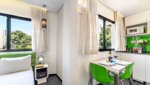 A modern hotel room with a bed, a desk with green chairs, a small kitchen area, and a view of greenery outside. - 130 Rock Apartments (Tel Aviv)