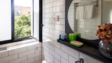 A bathroom with white tiled walls, a black countertop, a round mirror, a window, and a view of greenery outside.