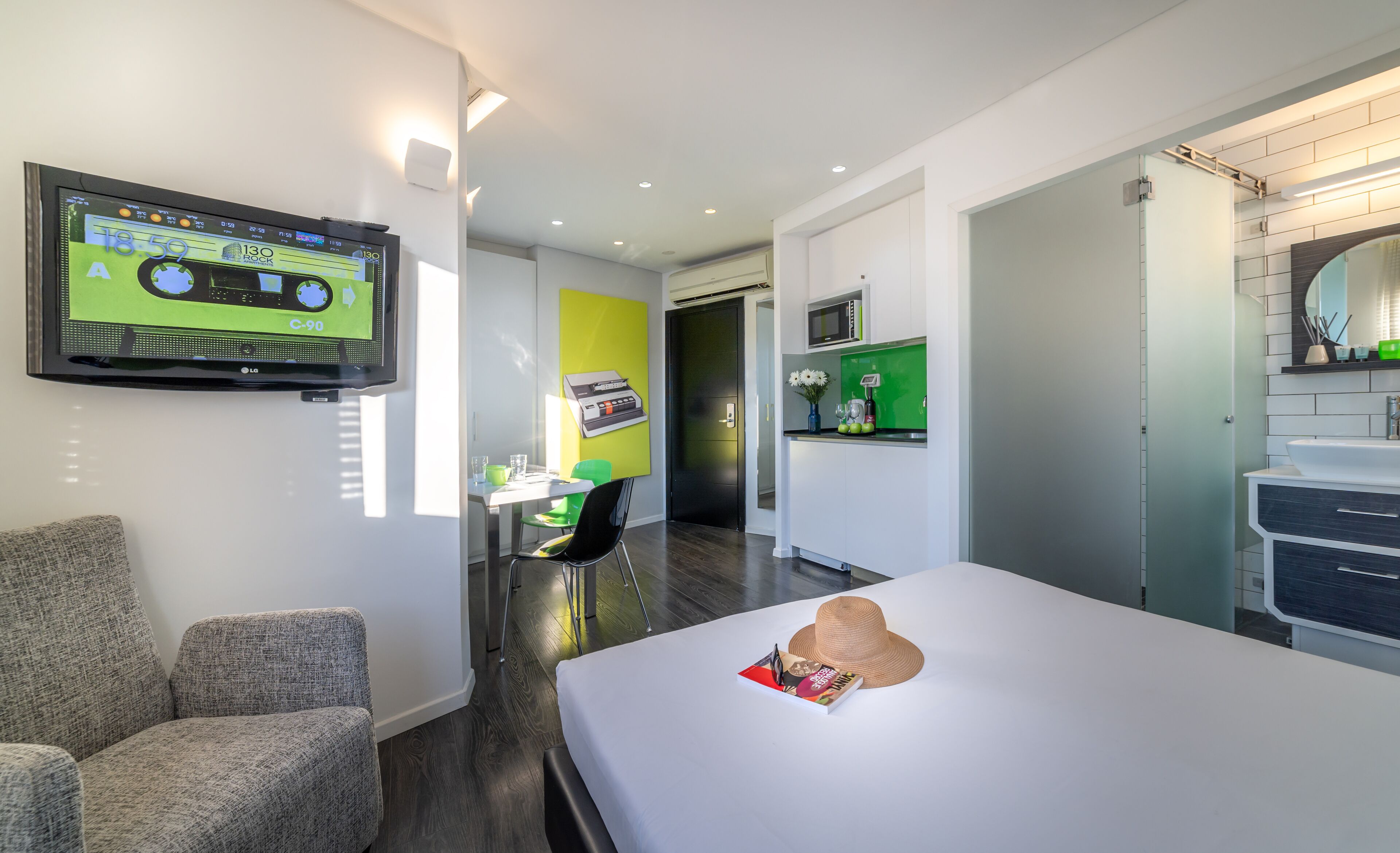A modern hotel room with a flat-screen TV mounted on the wall, a bed with a magazine and a hat on it, a small table with a chair, and a kitchenette area with a microwave and a sink.