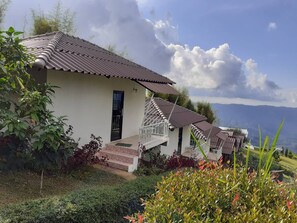 A row of white buildings with brown tiled roofs, a staircase leading to a balcony, and a view of mountains in the background.